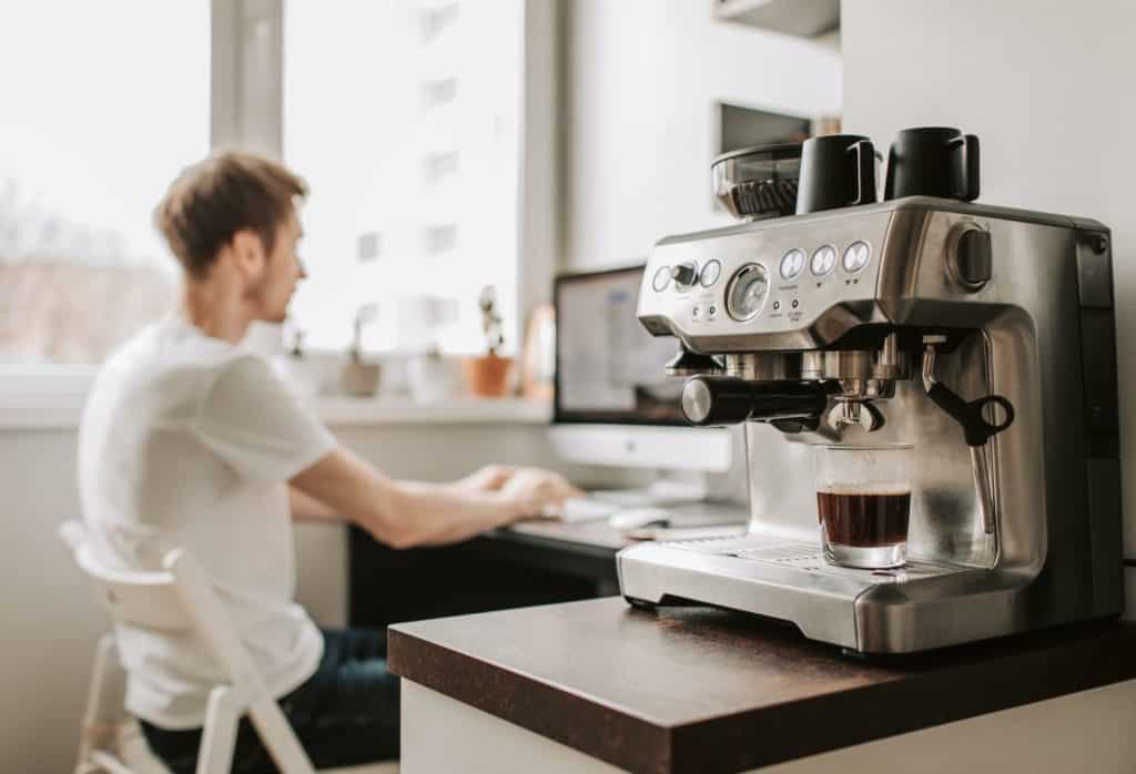 Bild zeigt eine Sieträgermaschine rechts im Vordergrund mit einem Glas Espresso unter dem Auslauf. Im Hintergrund ist ein junger Mann zu sehen, der auf einem weißen Klappstuhl sitzt und auf den Bildschirm eines Mac auf seinem Schreibtisch schaut.