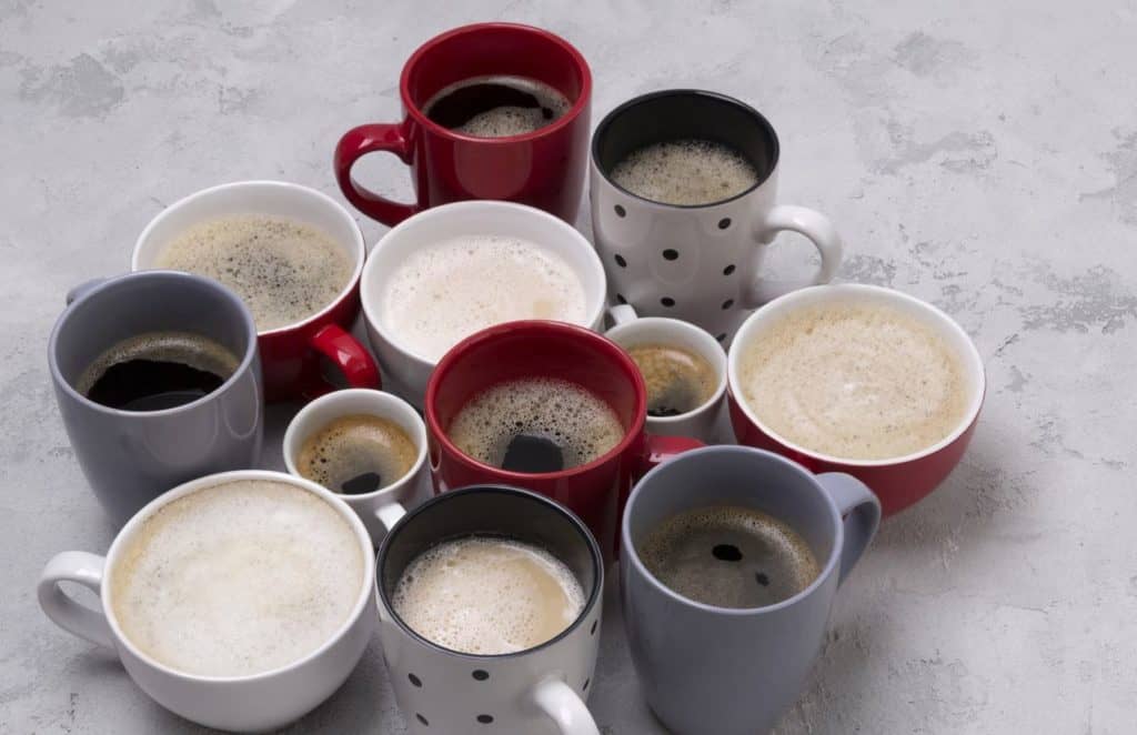 Mehrere Tassen in verschiedenen Formen, Farben und Größen stehen zusammen und sind mit unterschiedlichen Kaffeegetränken gefüllt.