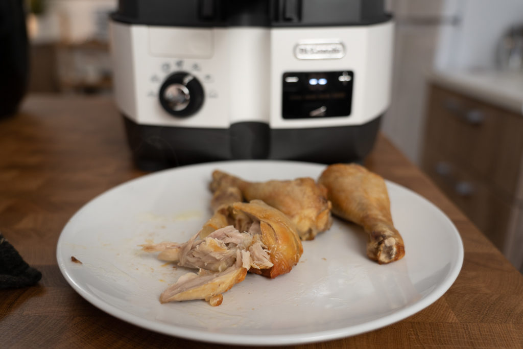 Drei fertig zubereitete Hähnchenschlegel liegen auf einem weißen Telle, während der untere Teil der DeLonghi MultiFry Extra Chef etwas unscharf im Hintergrund zu sehen ist