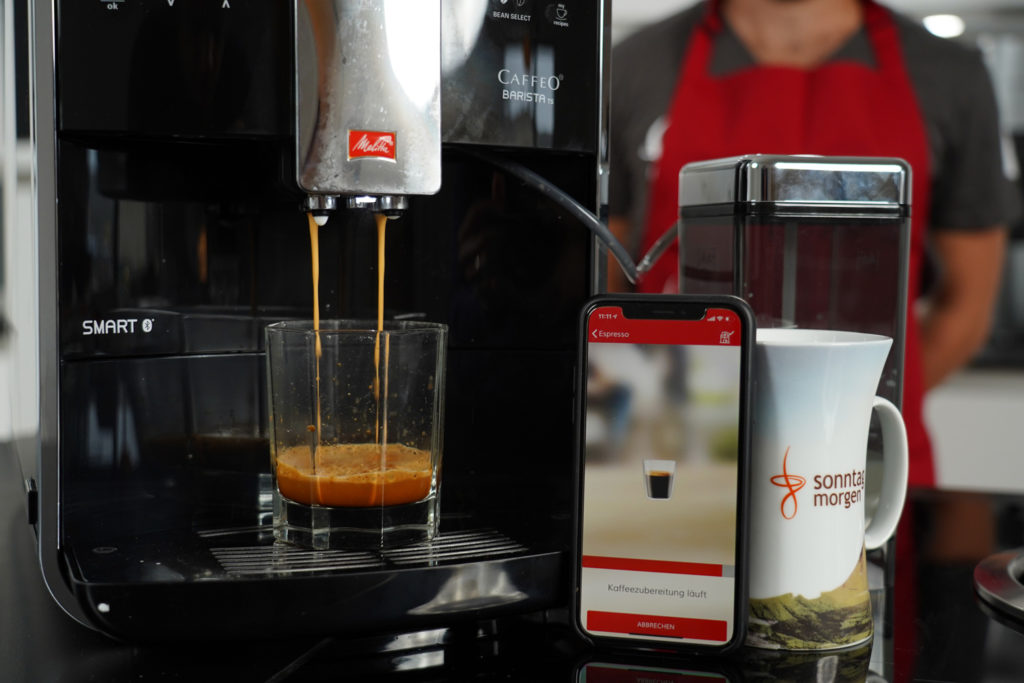 Melitta Caffeo Barista TS Smart - Espressobezug, rechts daneben ein Android Smartphone mit der Melitta-App, die den Verlauf anzeigt